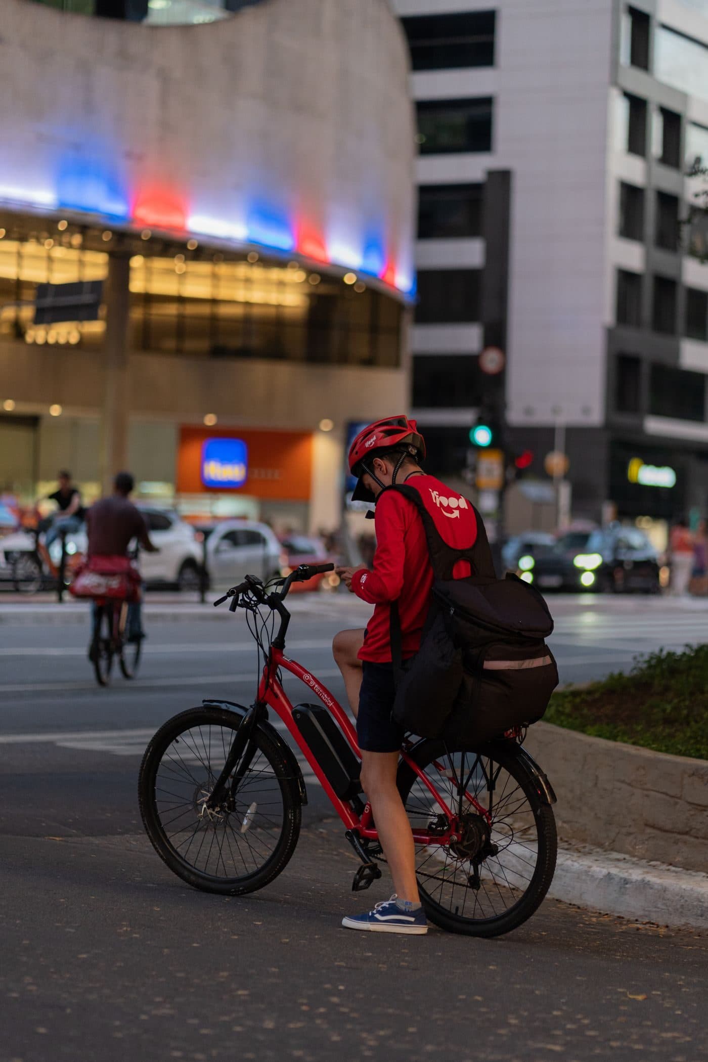 A delivery rider in a red jacket beside an e-bike on a city street at dusk
